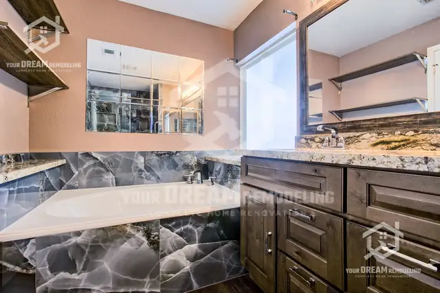Drop-in tub wrapped in dark marble-look tile beside an espresso vanity with open wall shelves
