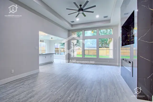 Open family room with gray wood-look plank flooring, tray ceiling, ceiling fan and a marble-look fireplace wall