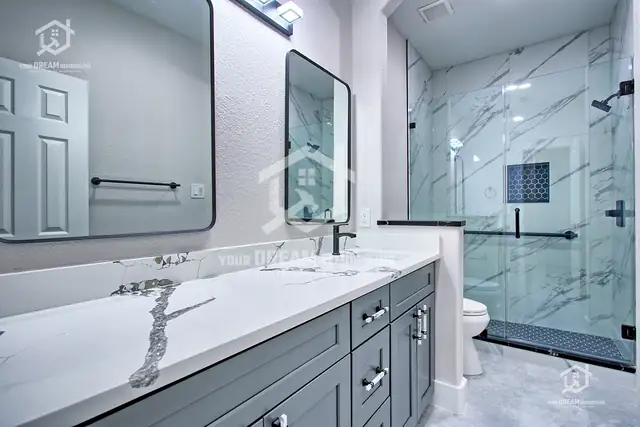 Bathroom with a long gray vanity, white quartz top, black faucets and a glass-enclosed shower with marble-look tile